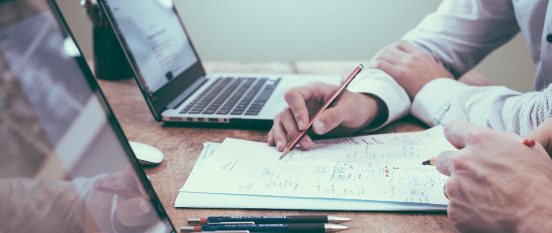 Dwie osoby analizujące dokument z odręcznymi notatkami i rysunkami technicznymi przy biurku z laptopem i ołówkami.