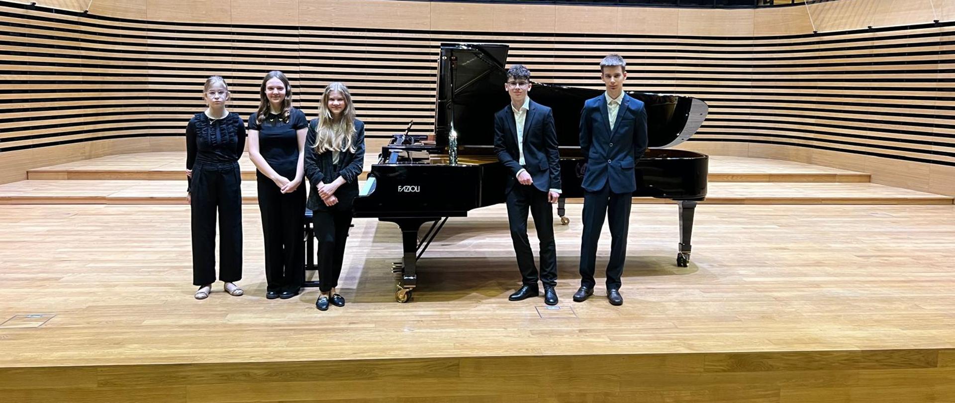 Trzy dziewczyny i dwóch chłopaków stoi na scenie przy fortepianie.