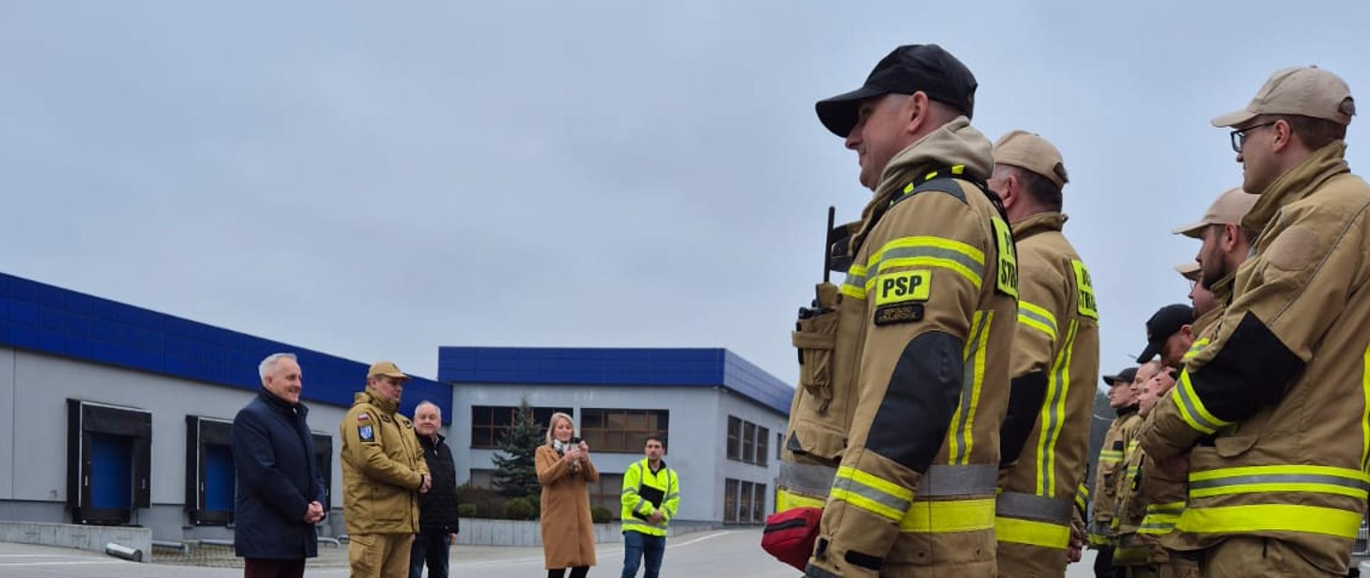 plac przed halą produkcyjną na pierwszym planie strażacy w ubraniach specjalnych stojący w dwuszeregu, naprzeciw nich komendant powiatowy państwowej straży pożarnej brygadier Marcin Mazurkiewicz, obok niego między innymi starosta sępoleński Jarosław Tadych.
