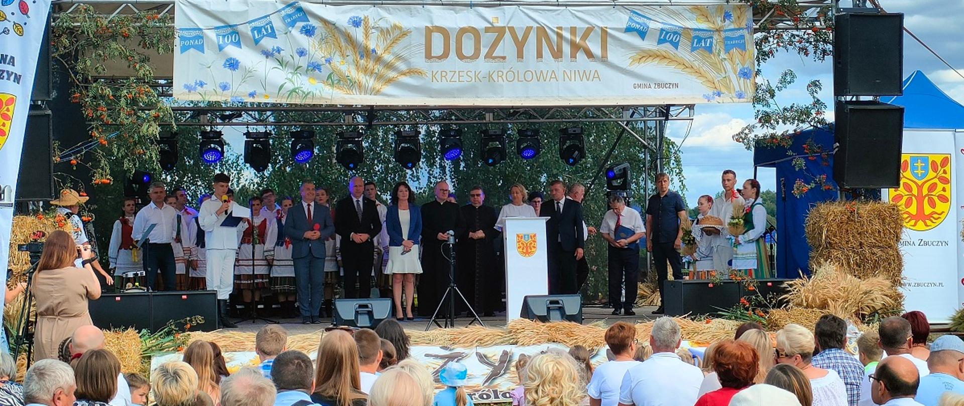 Dwadzieścia pięć osób stojących na scenie, część z nich ubrana jest w stroje ludowe. Stojący po prawej chłopiec trzyma bochen chleba, obok niego stoją dwie dziewczynki z palmami. Mężczyzna trzyma w ręce mikrofon i przemawia. Na górze sceny plakat z napisem Dożynki Krzesk Królowa Niwa. Z przodu sceny leżą snopki zboża, po bokach paczki słomy. Przed sceną siedzi tłum widzów. Z lewej strony sceny stoi kobieta robiąca zdjęcia osobom stojącym na scenie.