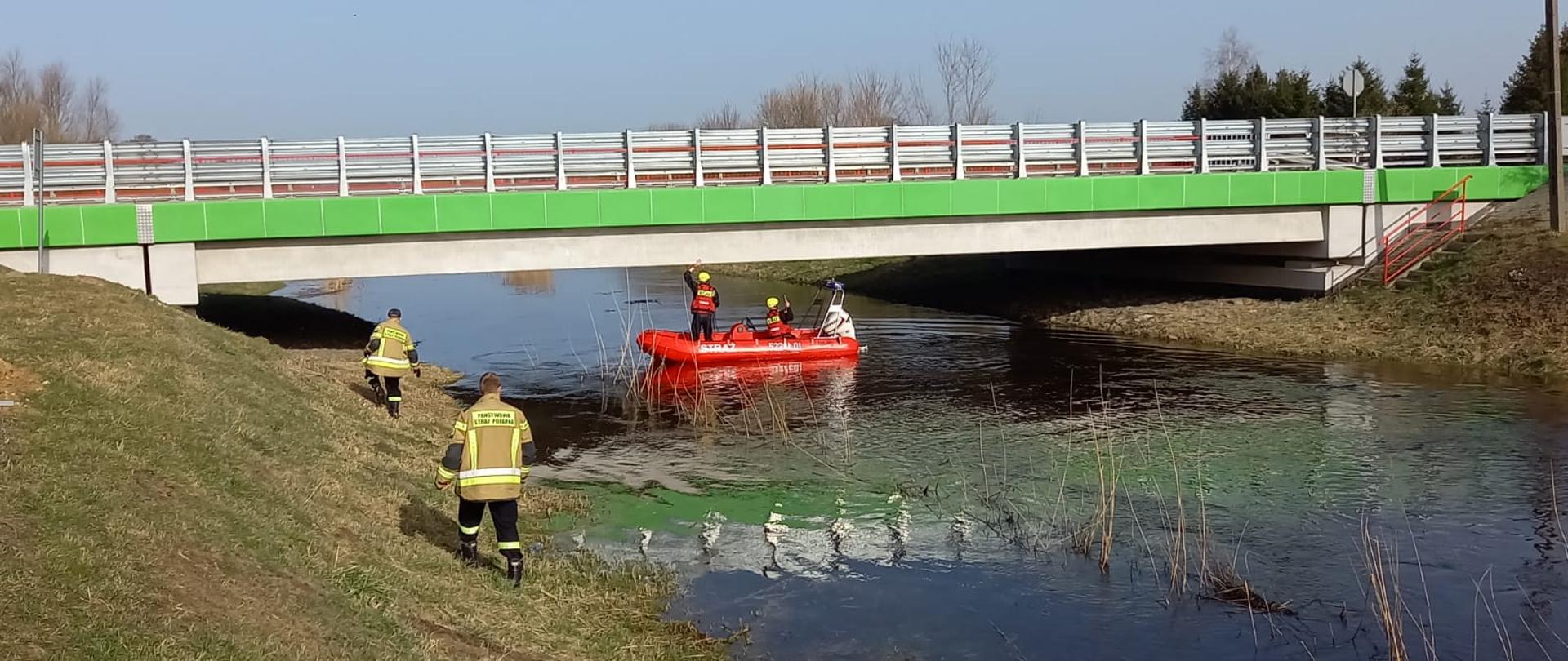 rzeka, nad nią zielono - biały most, pod którym przepływa ponton czerwony na nim strażacy patrolujący poziom rzeki