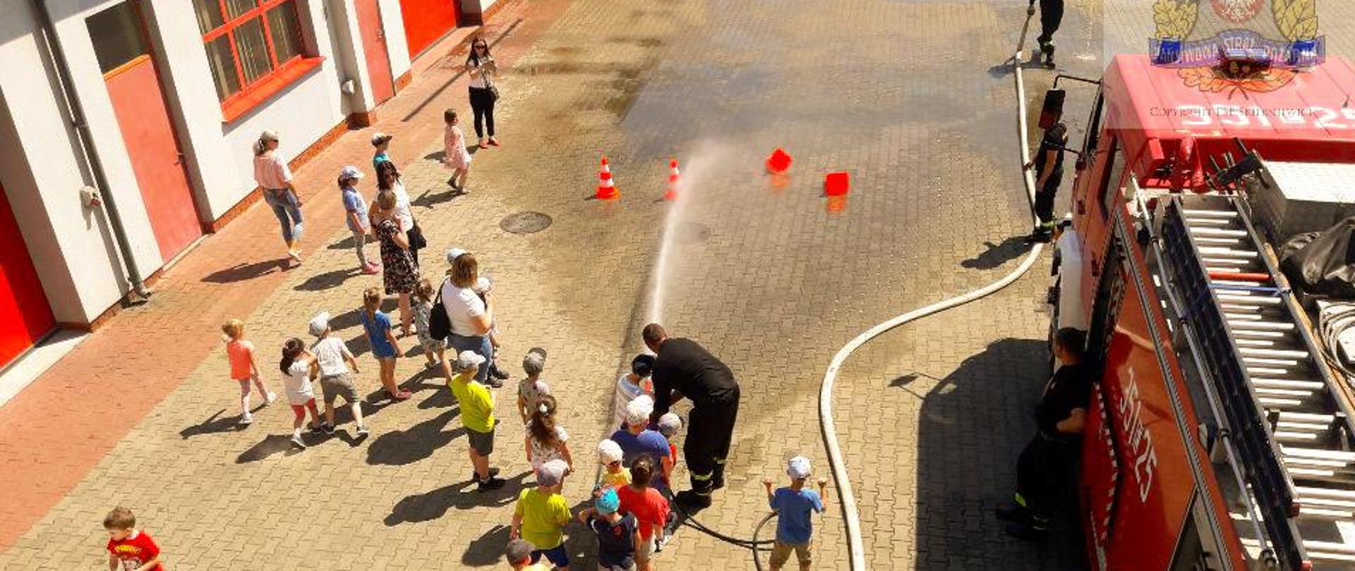 Zdjęcie przedstawia grupę dzieci podczas wizyty w siedzibie komendy miejskiej straży pożarnej w skierniewicach stojącą przed samochodem strażackim wraz ze strażakami pełniącymi tego dnia służbę. Dzieci uczestniczą w zabawie polegającej na przewracaniu pachołków za pomocą strumienia wody z węża strażackiego pod nadzorem strażaka.