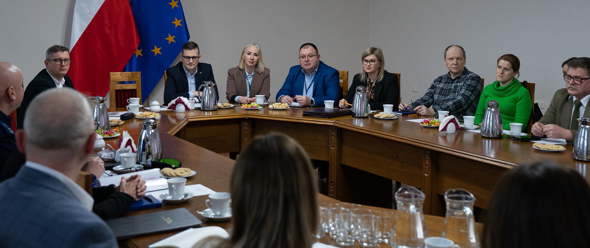 Na zdjęciu znajduje się grupa osób, która uczestniczy w spotkaniu dot. drogi ekspresowej s10. Na środku siedzi Wojewoda Kujawsko-Pomorski Michał Sztybel, a za nim stoi flaga polski i flaga unii europejskiej 