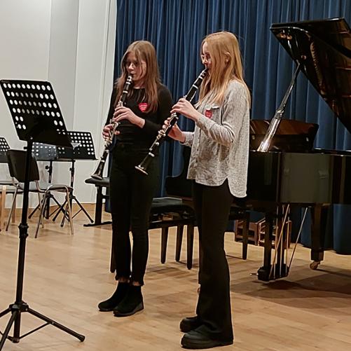 Zdjęcie przedstawia dwie klarnecistki grające na swych instrumentach z nut stojących przed nimi na pulpicie. Z tyłu po prawej stronie otwarty czarny fortepian. Z lewej strony pulpity i krzesła tworzące nieład. Uczennice ubrane są w czarne spodnie, jedna w białą druga w czarną koszulę. Włosy maja długie opadające na ramiona, na piersiach przyklejona naklejka z serduszkiem czerwonym WOŚP-u.