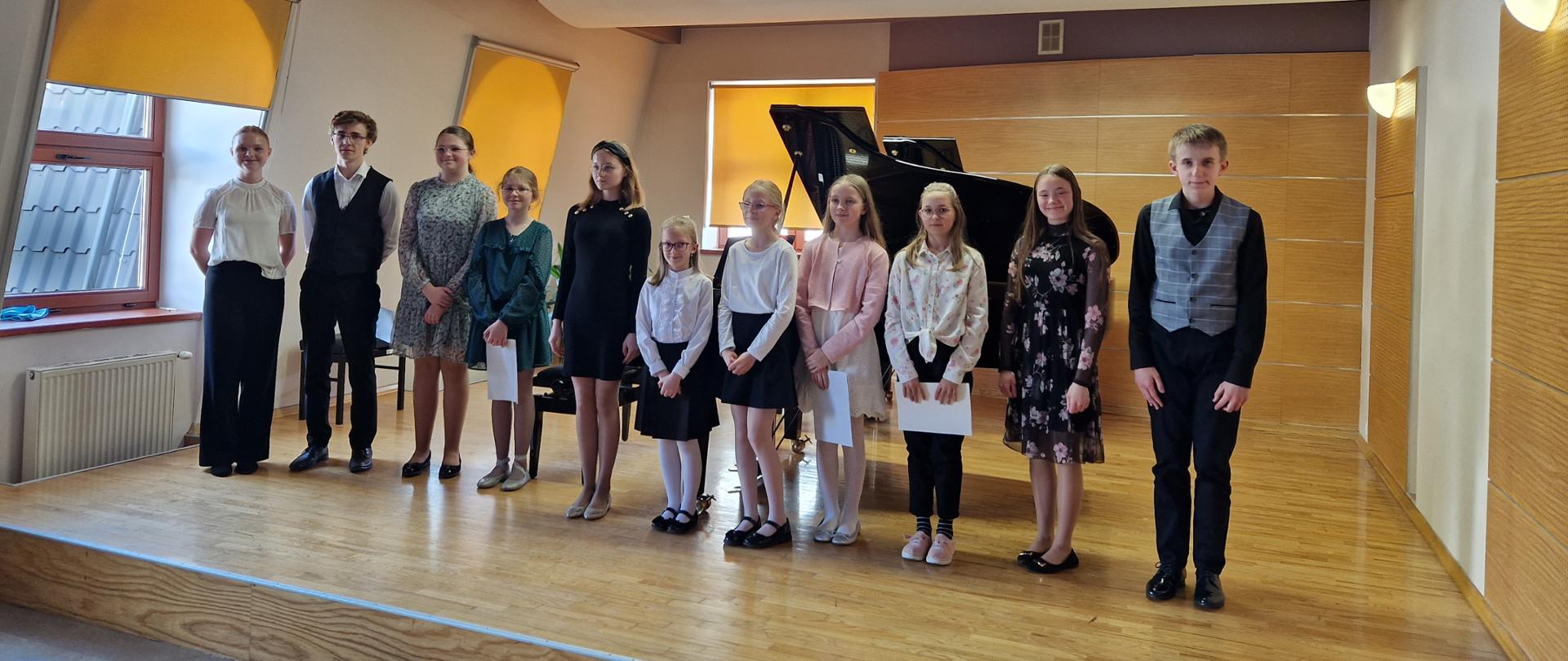 Wszyscy uczestnicy koncertu stoją na scenie sali kameralnej w jednym rzędzie, część uczniów trzyma w dłoni nuty. Za nimi dwa fortepiany oraz dwie ławy fortepianowe, po lewej stronie i z tyły za uczestnikami trzy okna przysłonięte roletami