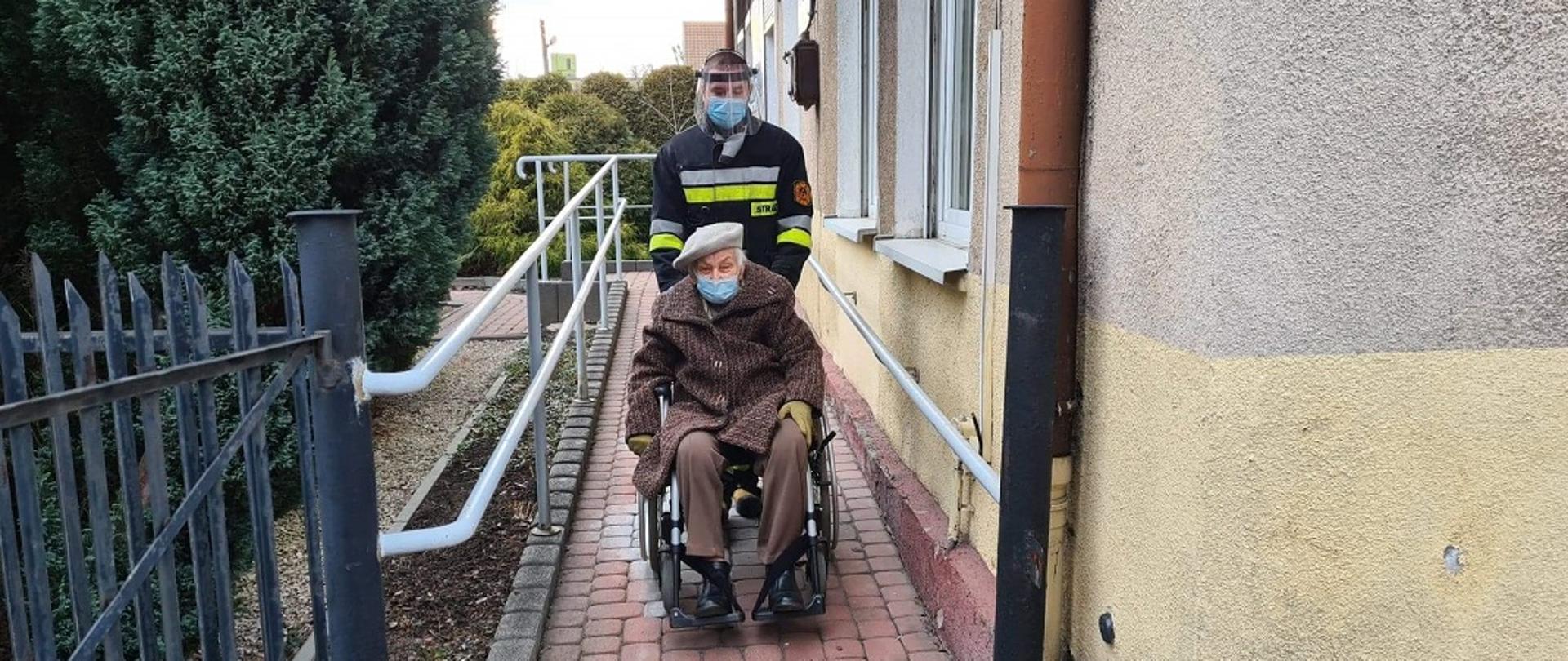 Na zdjęciu widać strażaka przed budynkiem, gdzie odbywają się szczepienia, który pcha wózek inwalidzki z kobietą udajacą się na szczepienie 