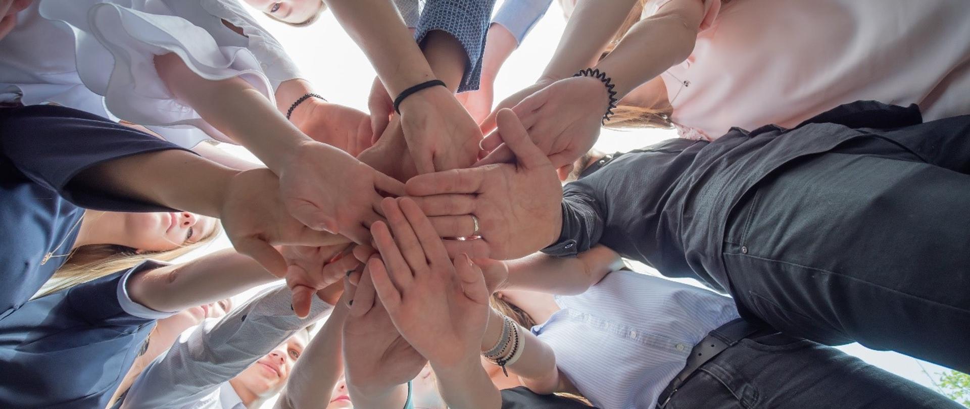 Na zdjęciu widać około 20 dłoni splecionych razem. Zdjęcie robione jest od dołu, mamy wrażenie, że leżymy na ziemi oglądając kilkanaście osób, którzy kładą dłoń na dłoni.