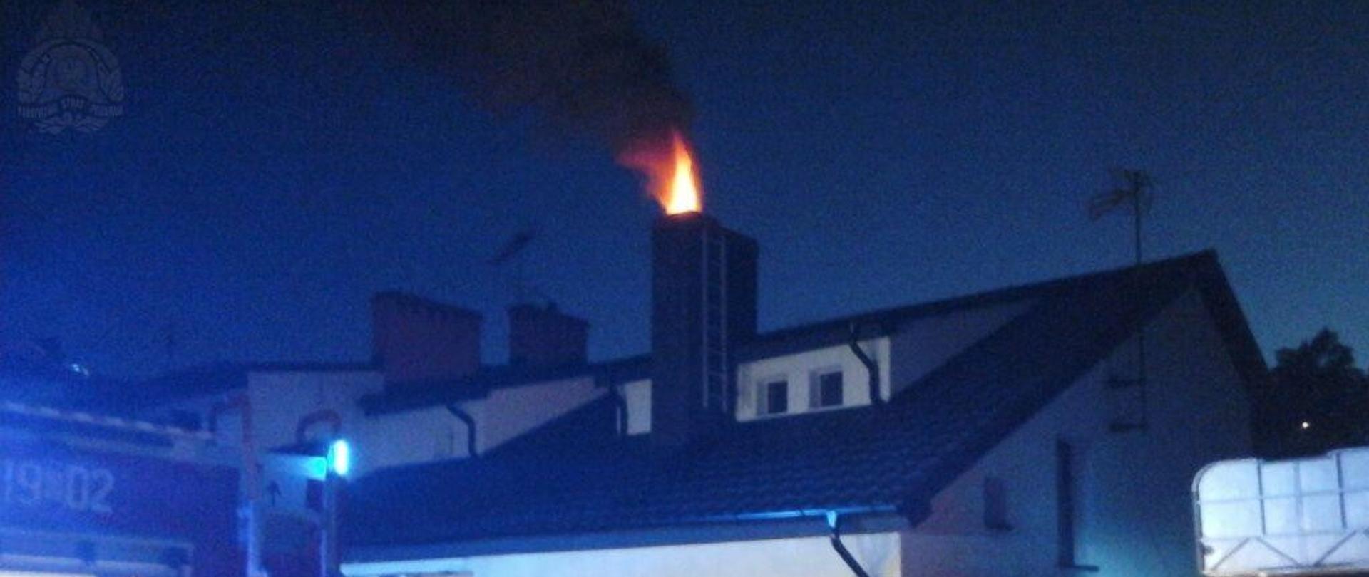 Z komina domu jednorodzinnego wydobywa się słup ognia. Wokół budynku strażacy prowadzą działania.