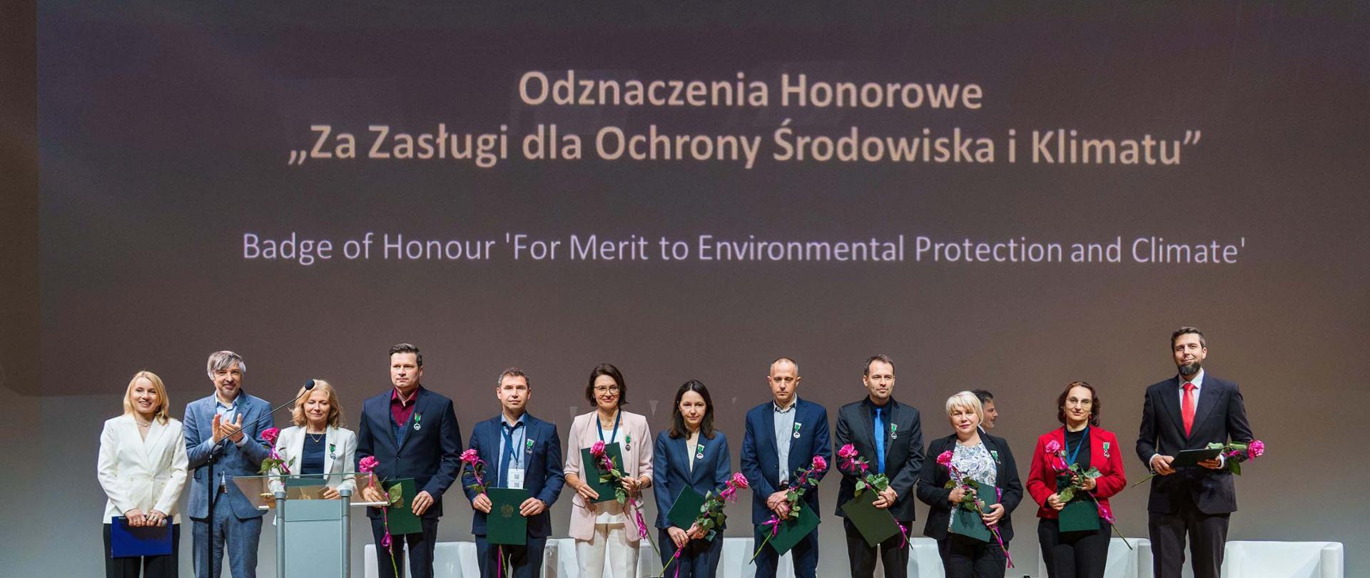 Sekretarz stanu w Ministerstwie Klimatu i Środowiska Krzysztof Bolesta stoi na scenie wśród ustawiony w szeregu osób odznaczonych za Zasługi dla Ochrony Środowiska i Klimatu. Odznaczeni, mężczyźni i kobiety, trzymają w dłoniach kwiaty i dyplomy. Wszyscy uśmiechają się. 