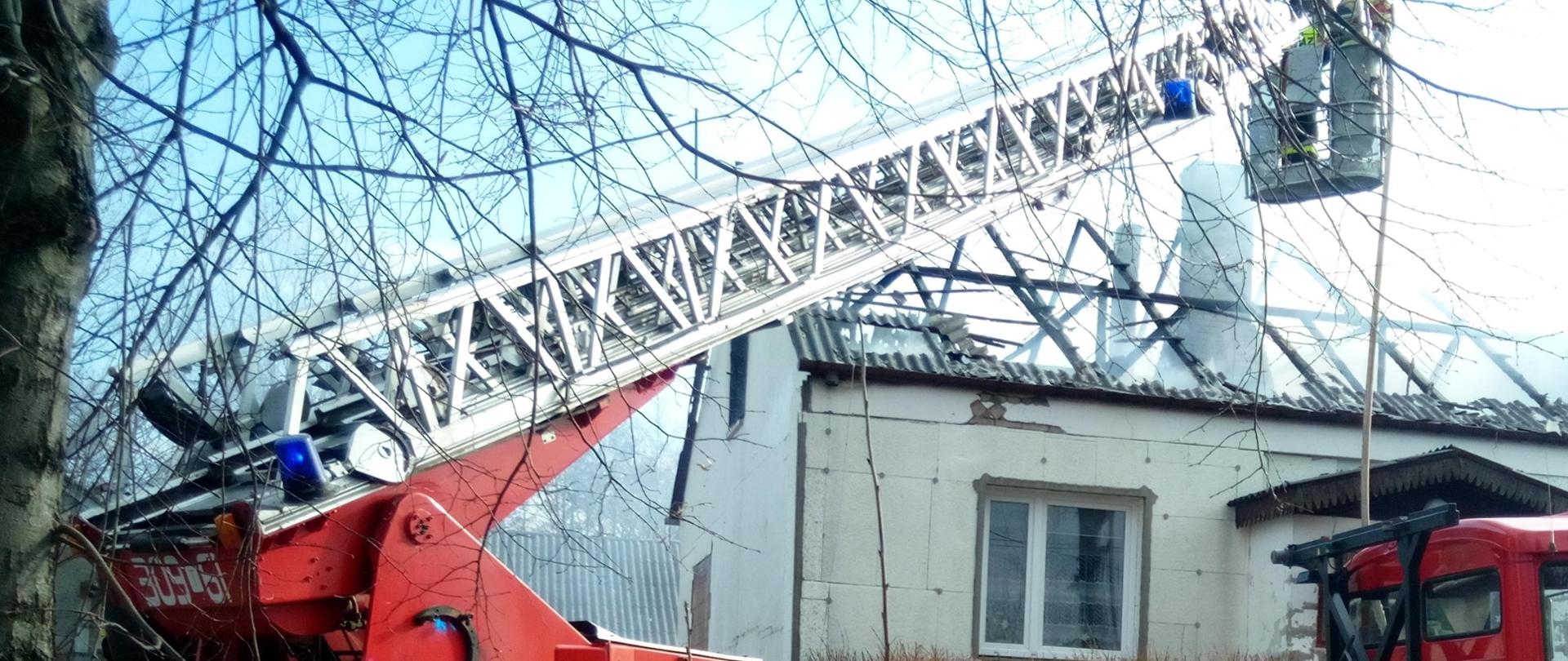 Na zdjęciu, na pierwszym planie widać samochód pożarniczy z drabiną mechaniczną. Za pojazdem znajduje się jednorodzinny dom, w którym wybuchł pożar.
