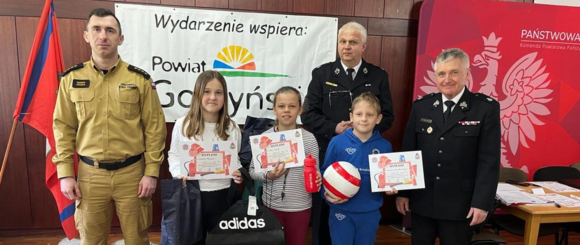 Zdjęcie przedstawia 2 dziewczyny oraz jednego chłopca, którzy zajęli miejsca od 1-3 w OTWP oraz Komendanta Powiatowego PSP w Gostyniu i 2 druhów OSP. Zwycięzcy trzymają w rękach nagrody oraz dyplomy.
