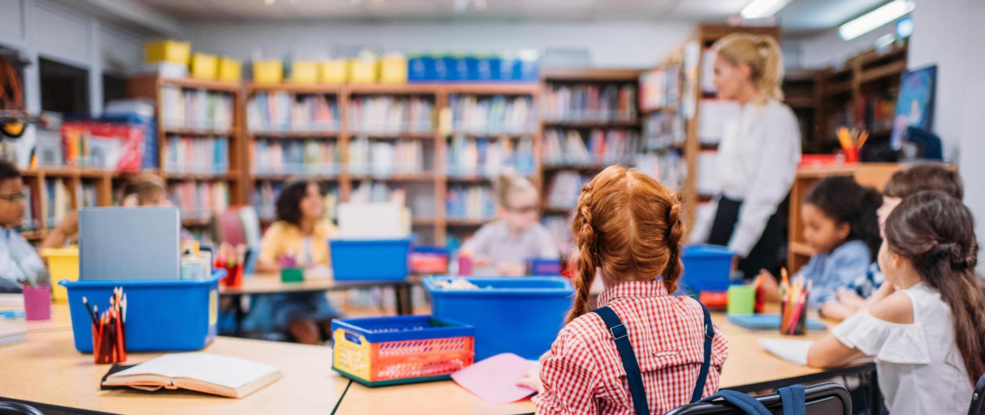Na zdjęciu jest nauczycielka wraz z dziećmi. Są w klasie lekcyjnej.