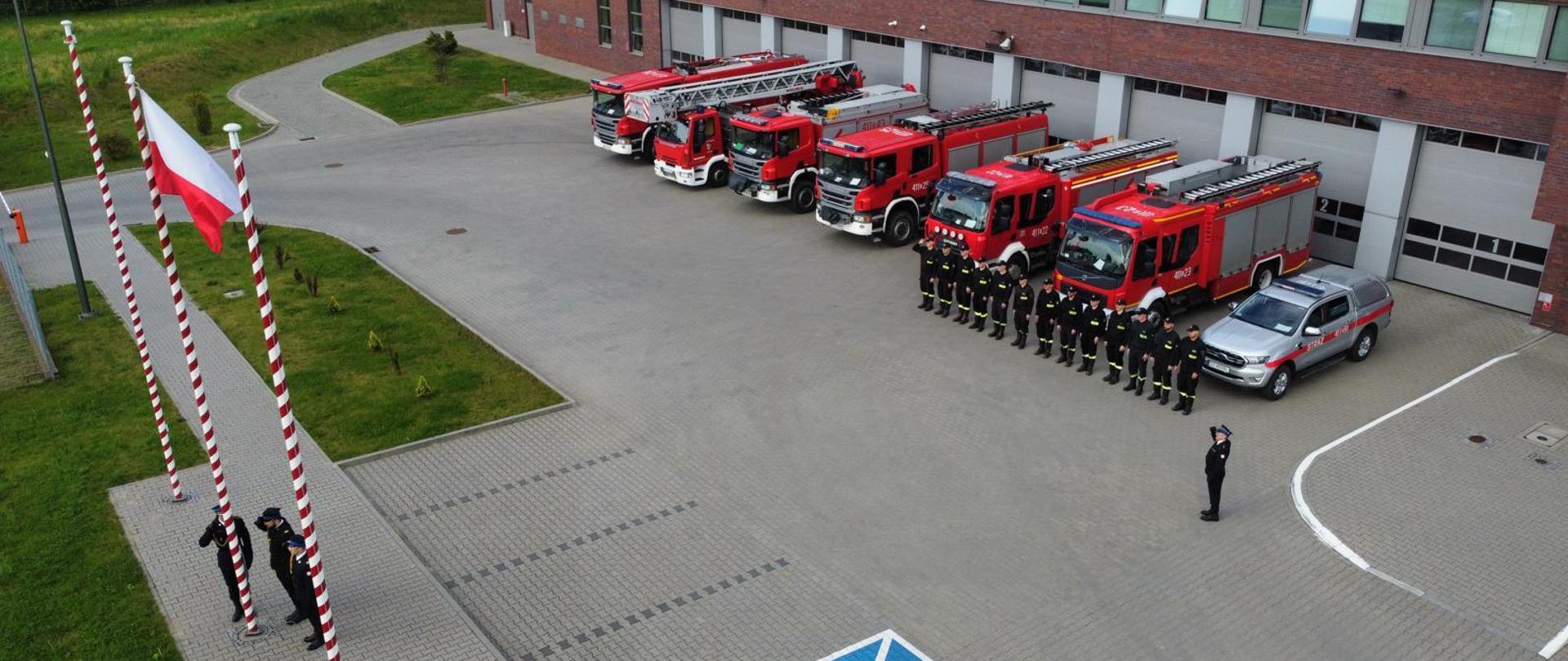 Na zdjęciu widoczny plac przed jednostką, z lewej strony poczet sztandarowy wciąga flagę na maszt. Z prawej strony strażacy ustawieni w szeregu, za strażakami ustawione wozy i budynek.
