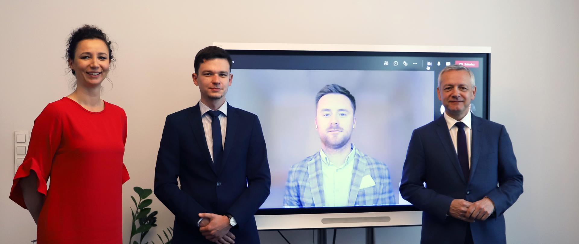 Zdjęcie - trzy osoby stoją przed ekranem do wideokonferencji, jedna widoczna na ekranie. Od lewej: Izabela Albrycht, Łukasz Gawron, na ekranie - Robert Siudak. Z prawej strony ekranu - minister Marek Zagórski.