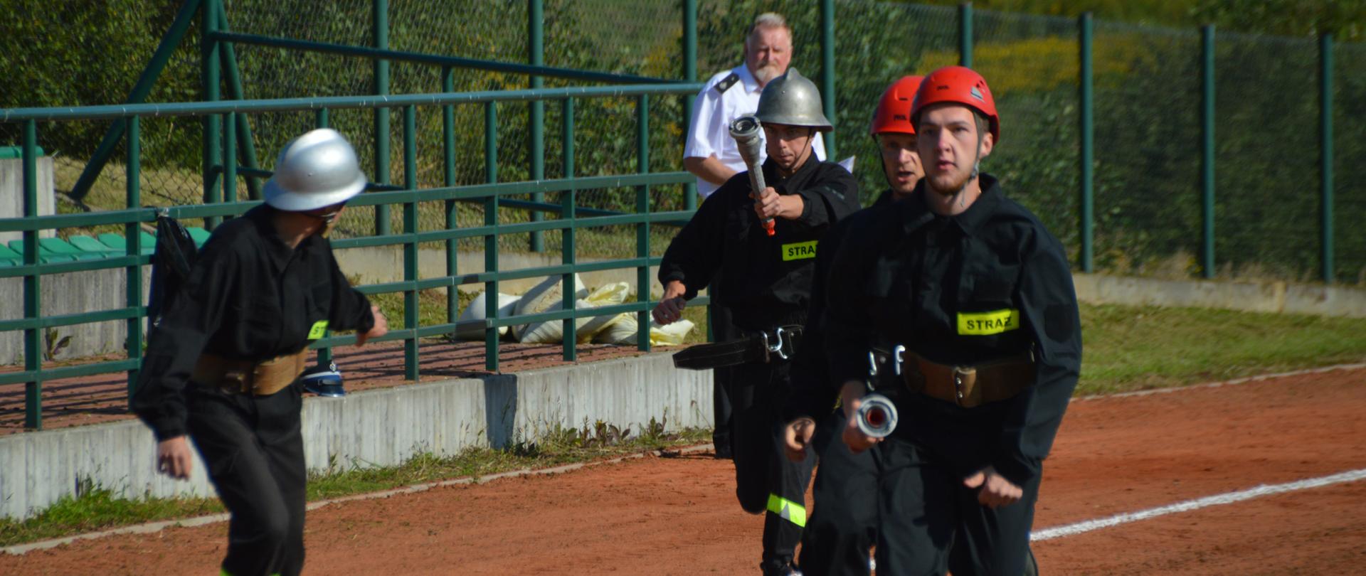 Na zdjęciu widać 4 strażaków rywalizujących w konkurencji bieg sztafetowy. Strażacy biegną z prądownicami trzymając je w prawych dłoniach biegną po bieżni koloru czerwonego. 2 ma hełmy kolory czerwonego, a dwóch koloru srebrnego. 