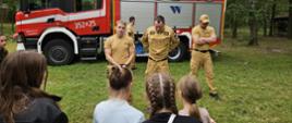 Na zdjęciu przedstawiono strażaków prowadzących zajęcia edukacyjne dla grupy dzieci i młodzieży w terenie leśnym. Spotkanie odbywa się przy samochodzie ratowniczo-gaśniczym, który stanowi element pokazu sprzętu wykorzystywanego w codziennej służbie. Strażacy prezentują wyposażenie pojazdu oraz omawiają zasady bezpieczeństwa, odpowiadając na pytania uczestników.