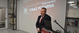 Przemawiający mężczyzna w garniturze, w tle czerwony slajd z rzutnika