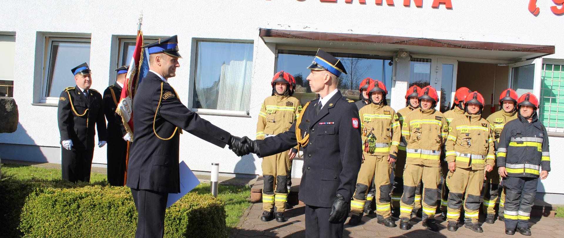 Zdjęcie przedstawia uroczystą zmianę służby Jednostki Ratowniczo-Gaśniczej w asyście Komendanta Powiatowego PSP w Świdwiniej, którzy znajdują się przy maszcie flagowym. Przed nimi znajdują się ustawione w dwuszeregu zmiana zdająca i przyjmująca służbę, ubrani w odzież specjalną z hełmami. W tle widać napis "Państwowa Straż Pożarna" na budynku komendy, drzewa. Podczas uroczystości jest wręczany akt mianowania na dowódcę JRG. dwóch strażaków jest ubranych w mundur galowy. Za mini znajduje się poczet sztandarowy.