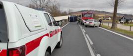 Na zdjęciu widać wypadek samochodu ciężarowego. Po lewej stronie zdjęcia widać biało czerwony pojazd straży pożarnej stojący na asfalcie. Po prawej stronie widać szary pojazd marki BMW jest to pojazd policji. Za samochodem cywilnym policji widać kolejny pojazd strażacki. Obok strażackiego pojazdu ciężkiego widać wywróconą ciężarówkę. 