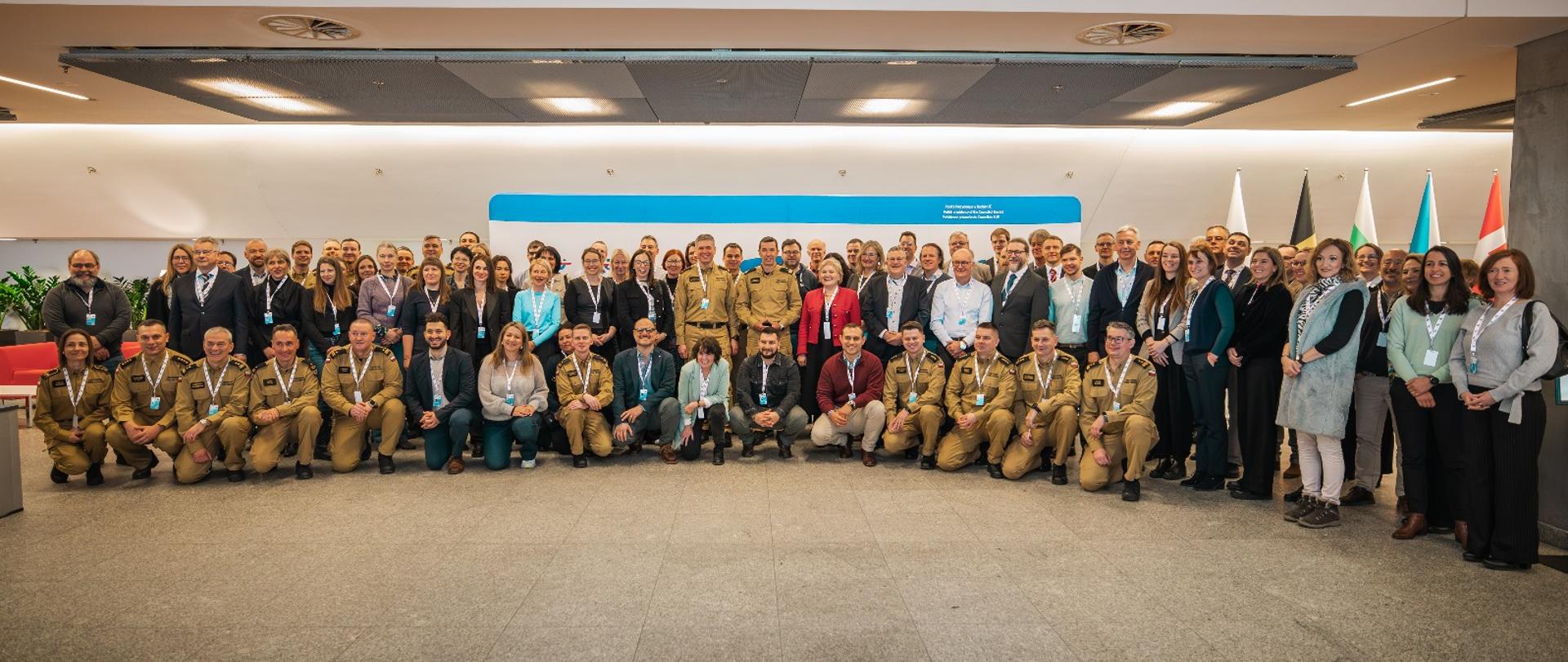 Zdjęcie grupowe kilkudziesięciu osób wewnątrz nowoczesnego budynku. Część osób nosi mundury w kolorze żółtym i kuca z przodu, a pozostałe osoby stoją za nimi. Wszyscy patrzą w kierunku aparatu. W tle widoczny jest znak z napisem oraz kilka flag. Na suficie znajduje się szereg lamp oświetlających przestrzeń.
