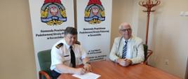 Podpisanie umowy na rozbudowę i przebudowę obiektów - brygadier Andrzej Wójtowicz, Komendant Powiatowy PSP w Szczecinku, siedzący przy stole z lewej strony ubrany w białą koszulę z czarnym krawatem popisuje umowę z siedzącym obok wykonawcą ubrany w beżową marynarkę, białą koszulę i krawat w biało-niebieskie paski.