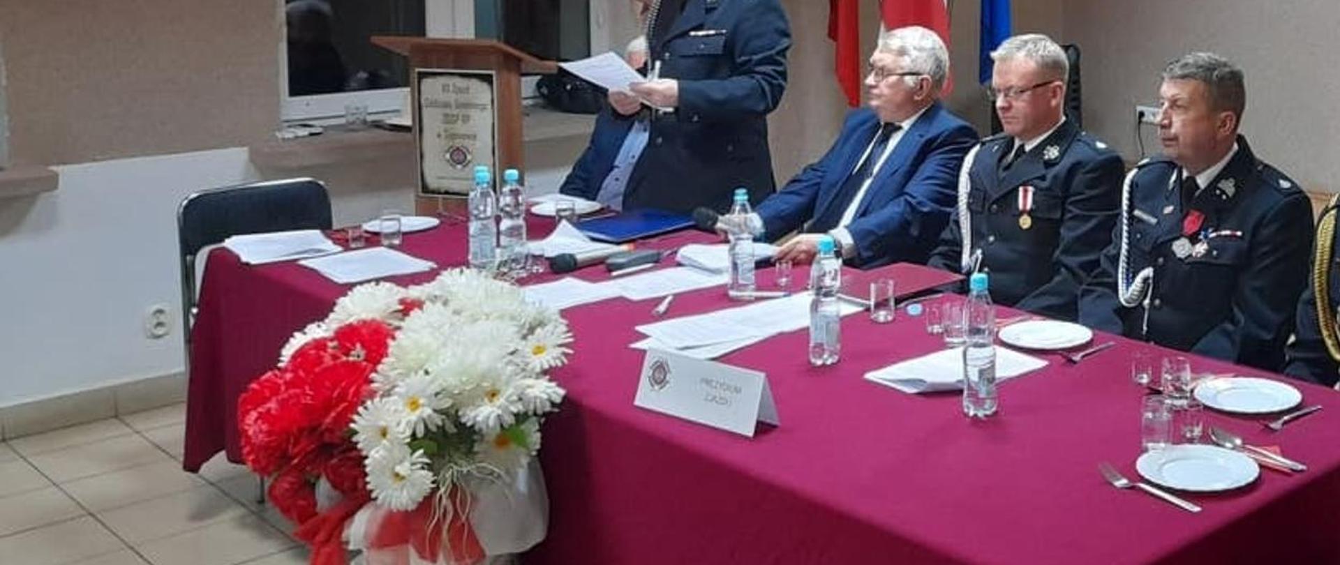 Strażacy oraz przedstawiciele samorządu siedzą przy stole. W tle godło oraz znaki ZOSP RP.