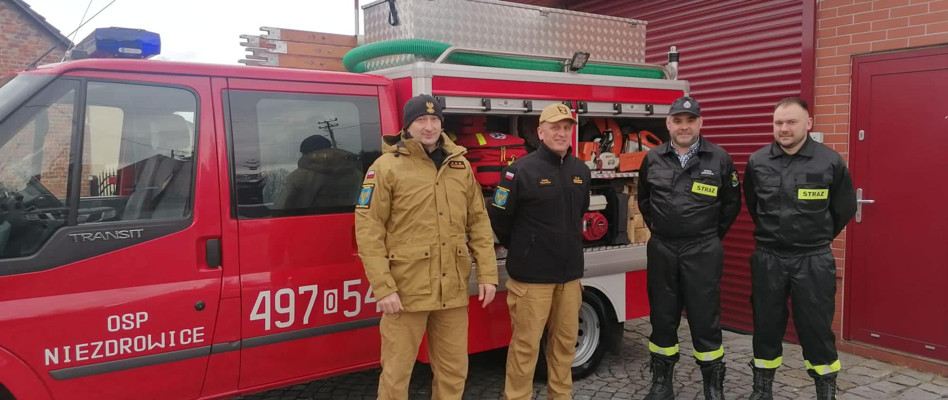 Na zdjęciu znajduje się samochód pożarniczy oraz czworo strażaków w mundurach