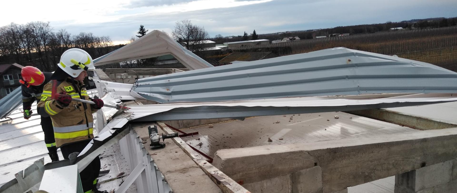 Na dachu budynku pracuje dwóch strażaków przy usuwaniu zerwanej i powyginanej blachy, która została uszkodzona w wyniku silnego wiatru. 