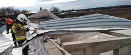 Na dachu budynku pracuje dwóch strażaków przy usuwaniu zerwanej i powyginanej blachy, która została uszkodzona w wyniku silnego wiatru. 