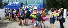 Na zdjęciu widocznych kilkunastu harcerzy pakujących bagaże do granatowego autobusu w związku z ewakuacją obozu harcerskiego. Harcerze stoją na szutrowej drodze za ich plecach plecaki karimaty i śpiwory.