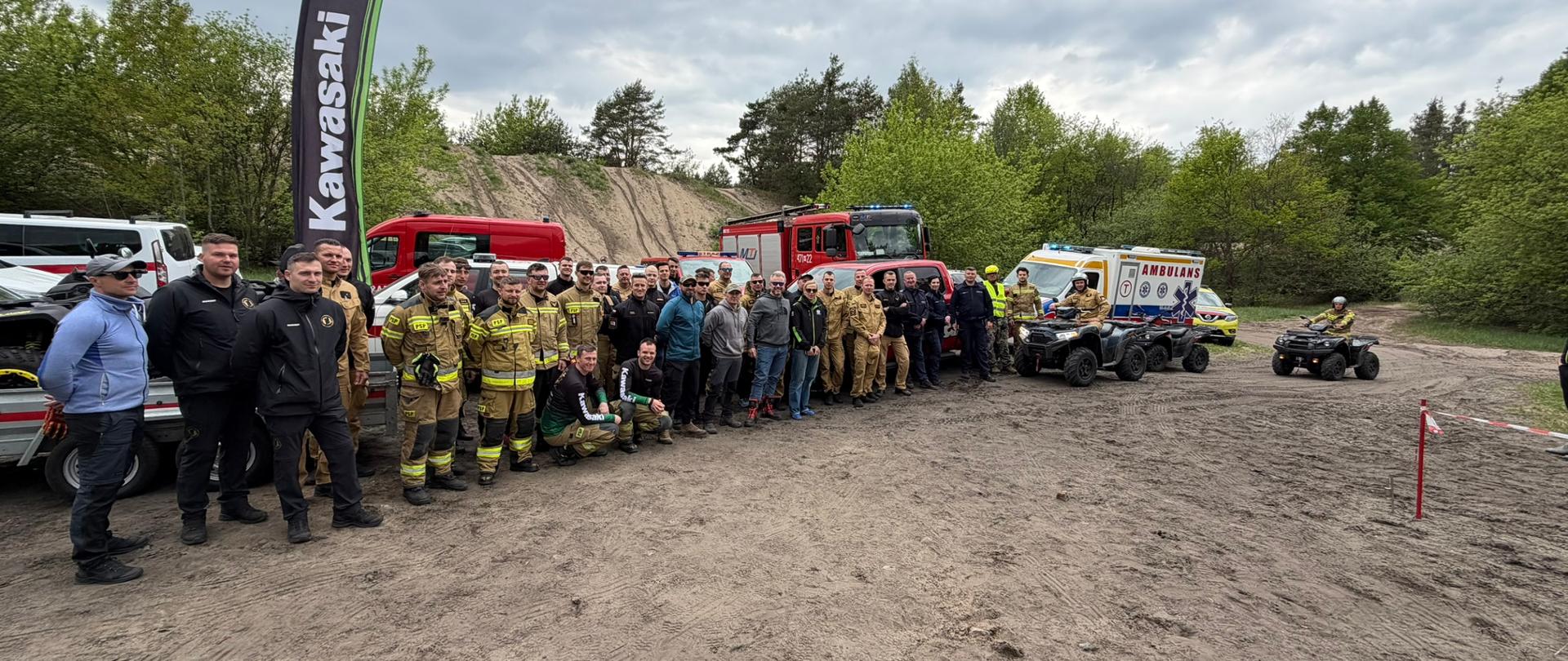 Uczestnicy szkolenia pozują do zdjęcia grupowego na terenie ćwiczeń. W tle znajdują się pojazdy ratownicze, karetka, quady, samochody pożarnicze oraz flaga z logo Kawasaki.

