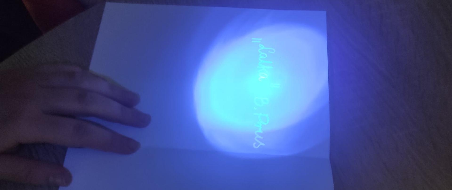 Zbliżenie na dłonie trzymające białą kartkę papieru w ciemniejszym pomieszczeniu. Światło latarki UV odsłania ukryty, fluorescencyjny napis: "Lalka" B. Prus.