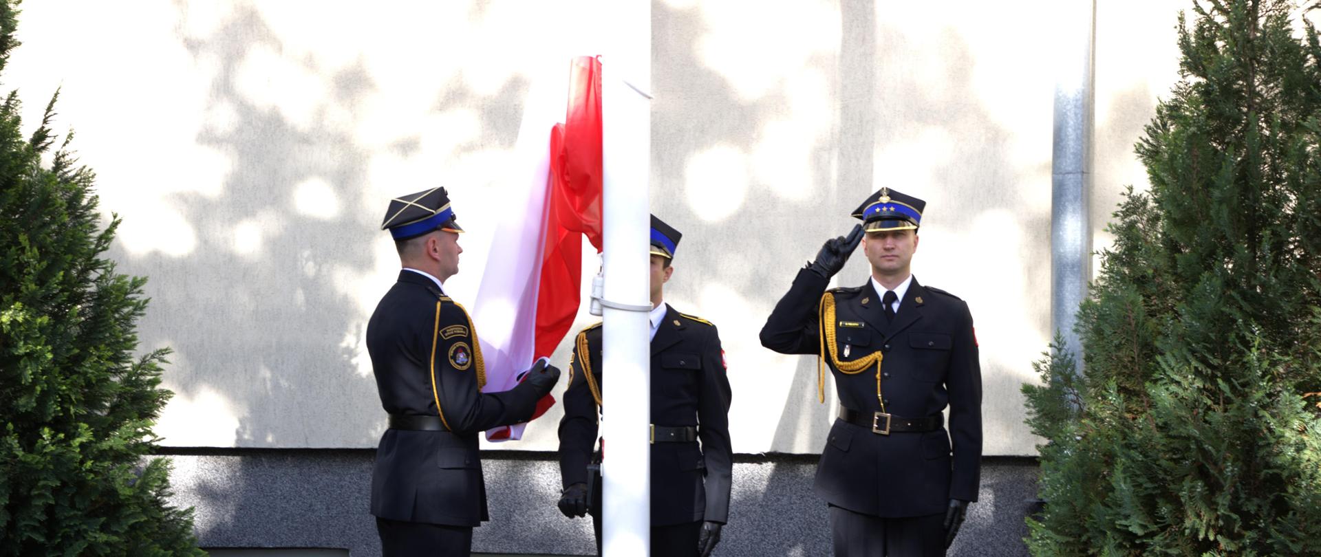 Trzech strażaków tworzących poczet flagowy stoi przy maszcie w celu podniesienia flagi państwowej. Jeden strażak podtrzymuje flagę państwową, drugi podnosi flagę na maszt, trzeci strażak stoi na baczność i salutuje.