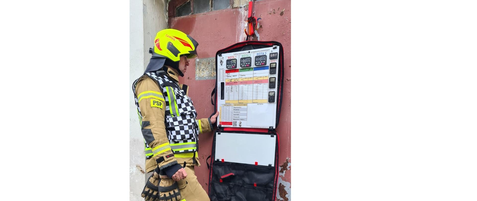 Strażak w żółtym hełmie i piaskowym ubraniu stoi przed tablicą na której są kolorowe tabele.