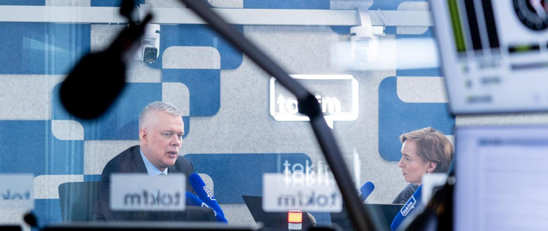 Na zdjęciu wnętrze studia radiowego. Przy stole, przy niebieskich mikrofonach z napisem TOK FM, siedzą dwie osoby - po lewej minister Tomasz Siemoniak, a po prawej redaktor Karolina Lewicka. Na pierwszym planie wiszący, czarny mikrofon radiowy. Po prawej stronie dwa monitory. Na dole zdjęcia mikser radiowy. U góry zdjęcia widać zegar, białe kamery oraz oświetlenie. Ściany w szaro-niebieskie wzory.