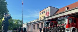 Strażacy świętują Dzień Flagi Rzeczypospolitej Polskiej. Na zdjęciu widać strażaków podczas uroczystego apelu z okazji Dnia Flagi, strażacy stoją w rzędzie, za nimi budynek strażnicy, flaga Polski uroczyście wciągana na maszt.