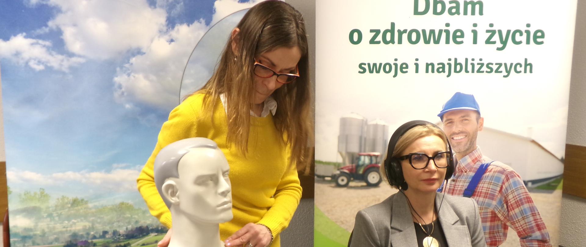 Pani Kierownik siedzi na krześle mając założone na uszach słuchawki. Obok niej znajduje się stół na którym są położone materiały i broszury dotyczące badania słuchu oraz pracownik firmy badającej słuch. Za Panią Kierownik znajduje się roll-up Kasy.