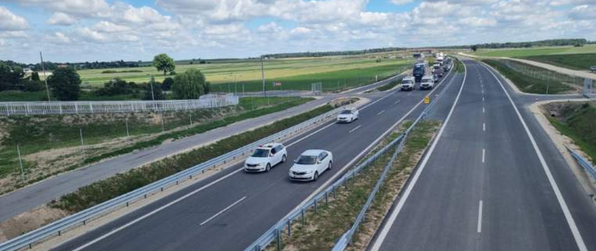 Nowa droga dwujezdniowa widziana z góry, z kilkoma samochodami jadącymi w jednym kierunku; w tle pola uprawne i błękitne niebo z rozproszonymi chmurami.