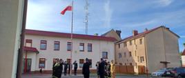 Uroczystość z okazji Dnia Flagi Rzeczypospolitej Polskiej. Strażacy ze zmian służbowych stoją naprzeciwko siebie. Poczet flagowy wciąga flagę państwową na maszt. Komendant, Zastępca Komendanta, poczet flagowy i dowódcy zmian salutują.