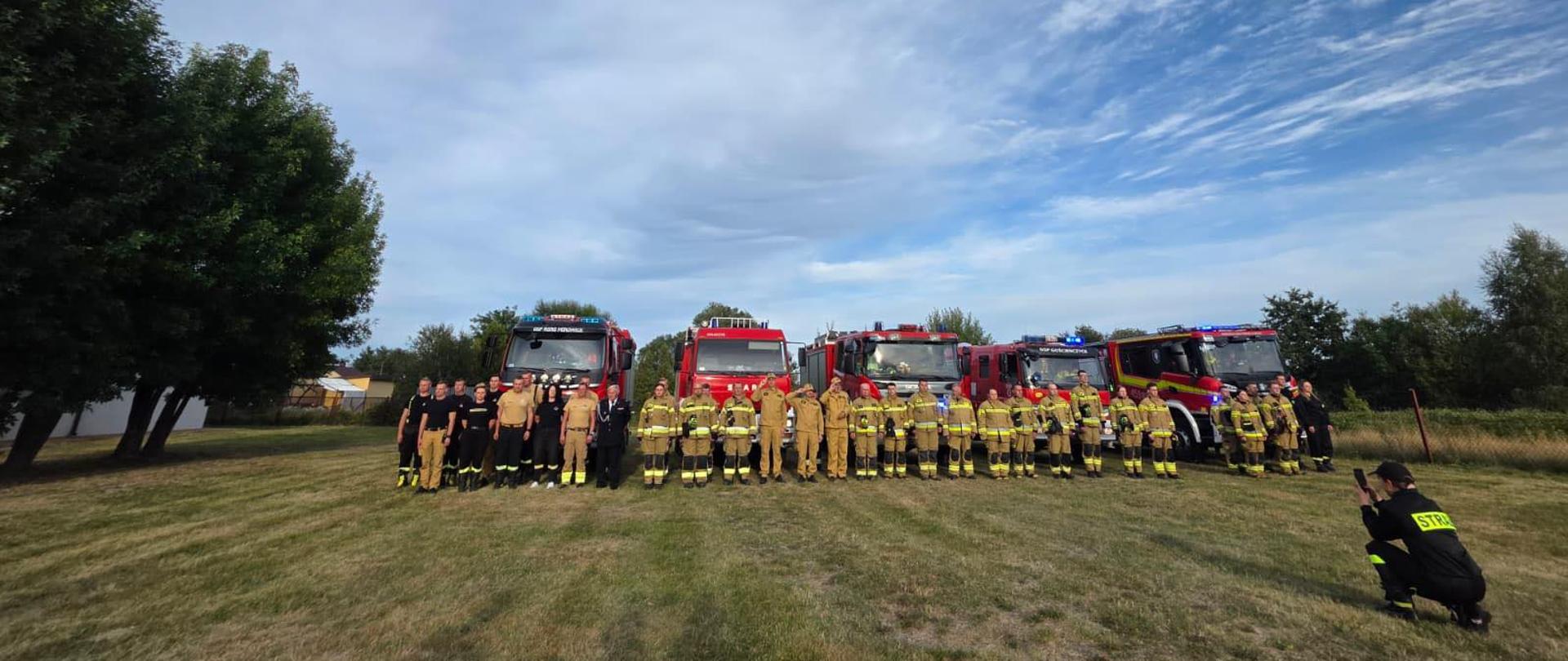 Na zdjęciu widoczna jest duża grupa strażaków stojących na trawie w równym szeregu. Za nimi ustawione są cztery duże, czerwone wozy strażackie z włączonymi światłami ostrzegawczymi. Po lewej stronie kadru rosną dwa duże drzewa, a po prawej widać ogrodzenie i więcej zieleni. Strażacy mają na sobie żółte i czarne stroje ochronne, część ubrana jest w czarne koszulki. Na pierwszym planie, z prawej strony, klęczy osoba w czarnym stroju z żółtym napisem „STRAŻ” na plecach i robi zdjęcie całej grupie. Nad sceną rozciąga się szerokie, niebieskie niebo z chmurami.