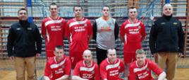 Na zdjęciu znajduje się grupa dziewięciu mężczyzn pozujących do wspólnej fotografii w hali sportowej. Stoją oni na tle bramki do piłki ręcznej lub futsalu z niebiesko-białym obramowaniem. Mężczyźni są podzieleni na dwa rzędy: Rząd tylny (stojący): Znajduje się tu sześciu mężczyzn. Osoby na skrajach (po lewej i prawej stronie) mundury służbowe w kolorze piaskowym. Na ich ramionach widoczne są pagony z dystynkcjami Państwowej Straży Pożarnej Trzej mężczyźni obok nich mają na sobie czerwone koszulki sportowe z białymi akcentami i napisem „KP PSP WOŁÓW”. Mężczyzna w środku rzędu nosi szarą koszulkę bramkarską z tym samym napisem. Rząd przedni (kucający): Czterech mężczyzn w identycznych czerwonych koszulkach sportowych i czerwonych spodenkach. Zdjęcie wykonane na parkiecie sportowym w kolorze jasnego drewna z widocznymi czerwonymi liniami boiska. Za bramką znajduje się ściana z drewnianymi drabinkami gimnastycznymi.
