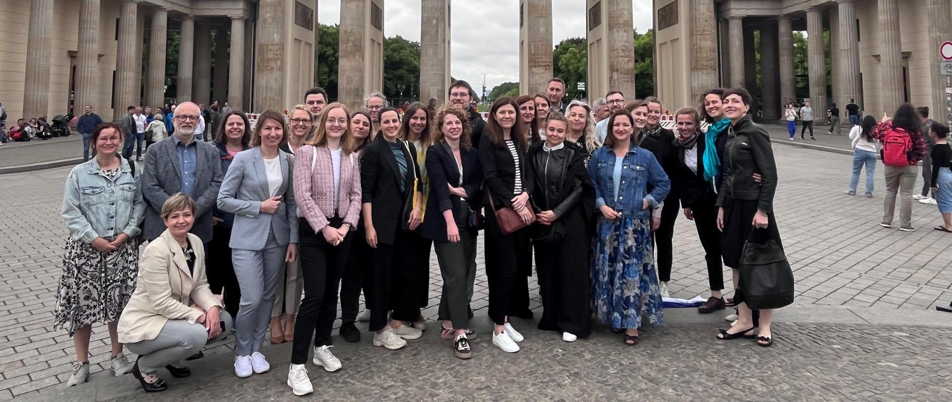 Na tle Bramy Brandenburskiej stoi duża grupa uśmiechniętych kobiet i mężczyzn. 