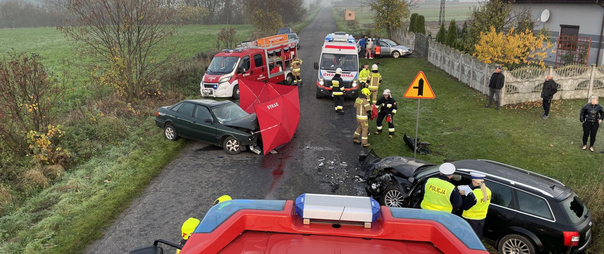 widok z góry zdarzenia na którym znajduję się czarny samochód z rozbitym przodem przy nim stoją Policjanci oraz drugi samochód koloru zielonego przed nim czerwony parawan za nim karetka pogotowia oraz inny samochód strażaki. Na poboczu stoją osoby postronne z boku widoczny budynek mieszkalny.