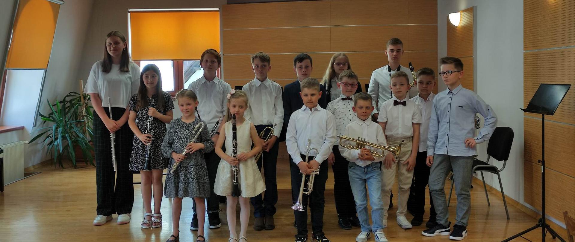 Wszyscy uczestnicy koncertu stoją w dwóch rzędach w sali kameralnej na scenie. Trzymają w dłoniach swoje instrumenty. Za nimi fortepian, dwa okna przysłonięte w części roletami, pod jednym oknem kaloryfer, oraz palma doniczkowa. Z prawej strony jedno puste krzesło i jeden pulpit.