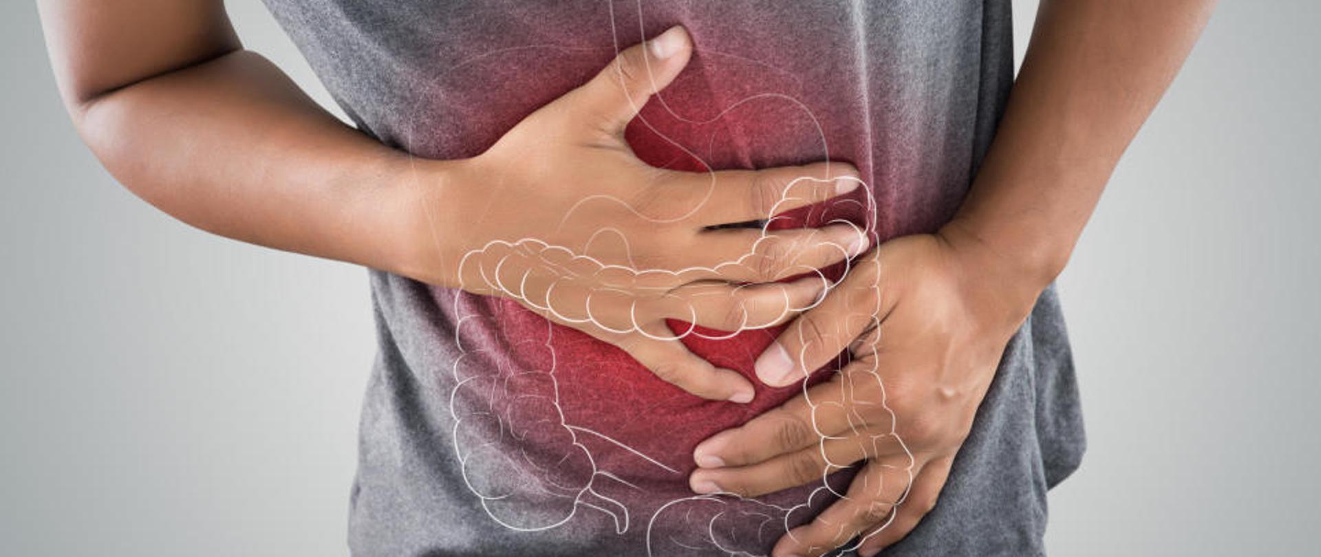 Na zdjęciu widoczne ręce trzymające brzuch. Na środku obrazka widoczny znak wodny jelit i żołądka. Siwa koszulka na brzuchu w kolorze czerwonym symbolizuje stan zapalny brzucha. Postać wygięta do przodu na znak boleści brzucha.