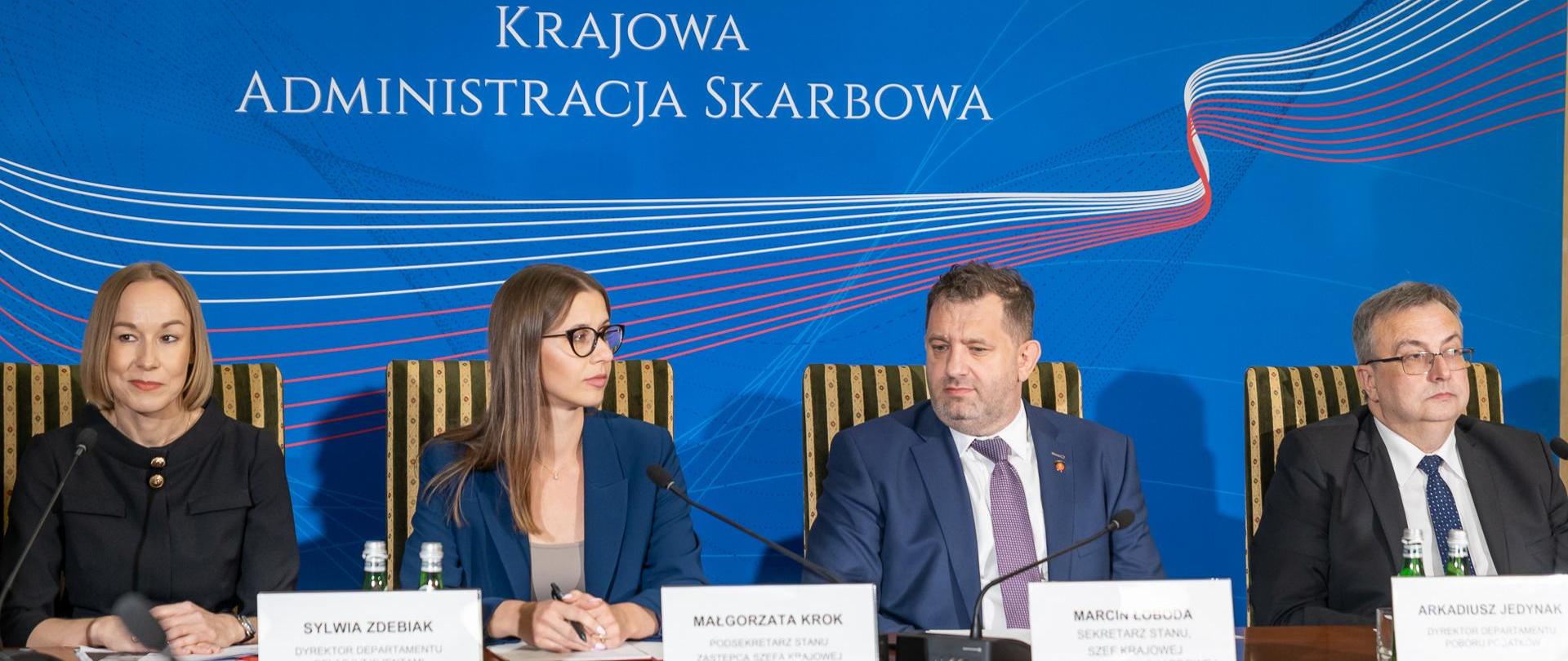 Szef KAS Marcin Łoboda, oraz zastępca szefa KAS Małgorzata Krok wraz z innymi prelegentami na konferencji prasowej w tle ścianka z napisem Krajowa Administracja Skarbowa