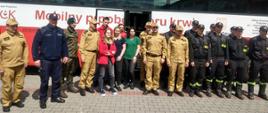 Na zdjęciu widać strażaków w ubraniach koszarowych w kolorze czarnym i piaskowym, wraz z nimi są funkcjonariusz Policji w granatowym umundurowaniu oraz osoba w mundurze wojskowym a także osoby cywilne. Wszyscy stoją na tle dużego autobusu w kolorze biało – czerwonym z napisem „Mobilny punkt poboru krwi”