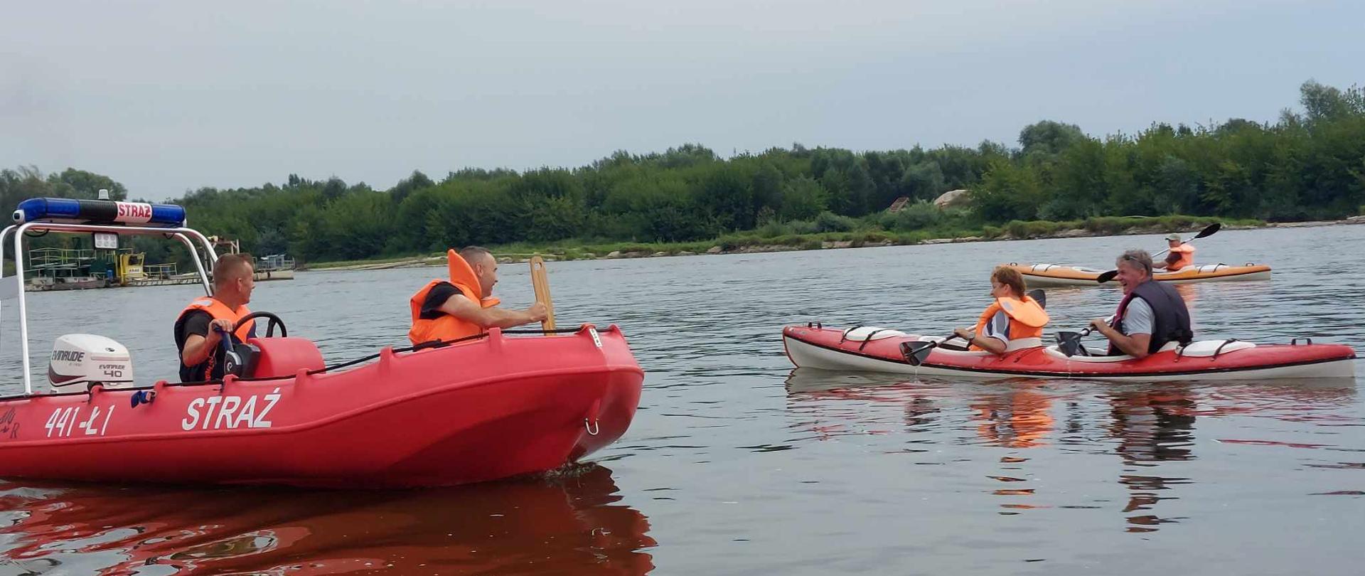 Zdjęcie przedstawia dwóch funkcjonariuszy PSP na łodzi motorowej na rzece Wisła.. Obok łodzi motorowej płyną dwa kajaki z turystami. w tle rzeka Wisła, drzewa oraz woda 