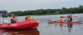 Zdjęcie przedstawia dwóch funkcjonariuszy PSP na łodzi motorowej na rzece Wisła.. Obok łodzi motorowej płyną dwa kajaki z turystami. w tle rzeka Wisła, drzewa oraz woda 