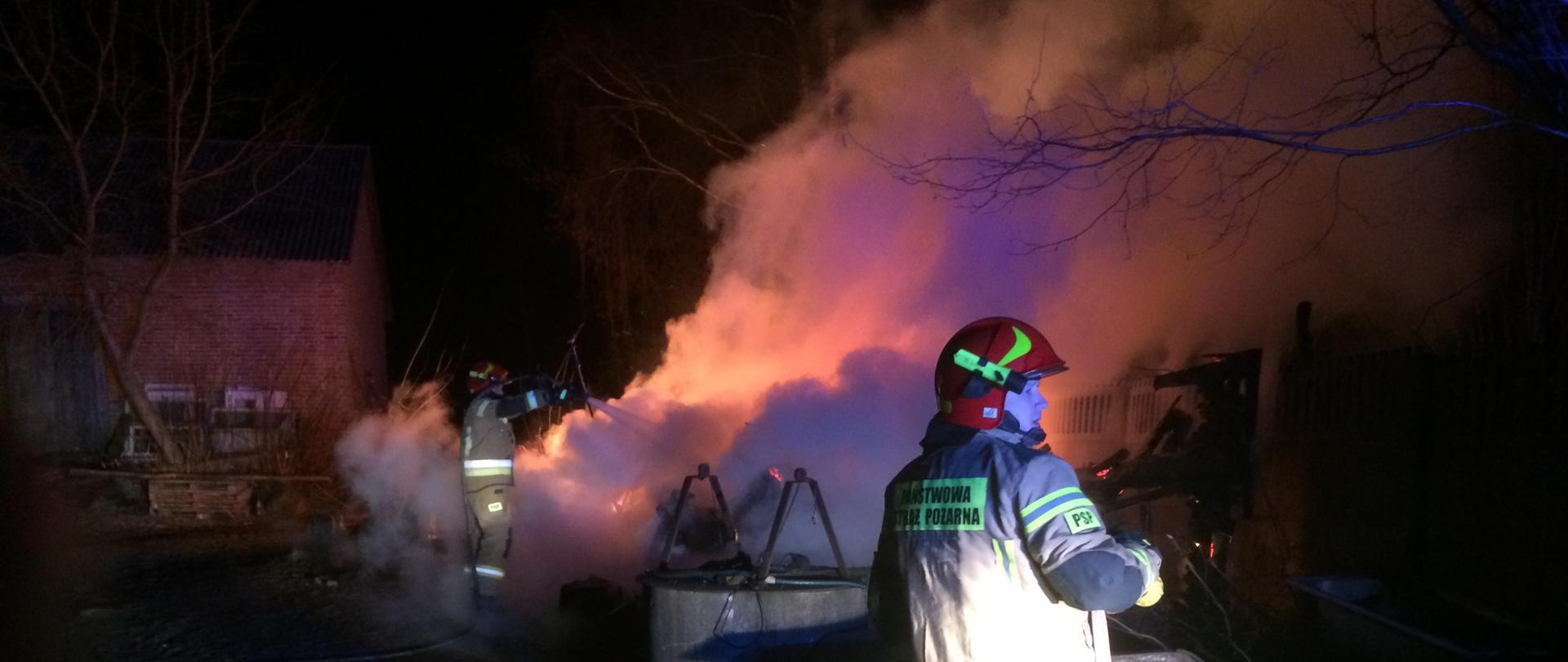 Zdjęcie zrobione jest w nocy. Na pierwszym planie znajduje się strażak w pisakowym ubraniu strażackim z hełmem na głowie, stojący tyłem do fotografa ,patrzący w prawo, w ręku trzyma wąż strażacki. Na drugim planie widać podwórko gospodarstwa na którym wybuch pożar szopy. Widać ogień oraz drugiego strażaka gaszącego pożar.